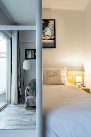 A hotel room with a bed, a nightstand with a lamp, a chair, a window with curtains, and a painting on the wall. - Le Royal Studio adorable avec terrasse (Cannes)