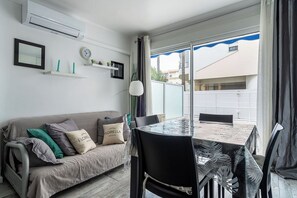 A modern living room with a sofa, a glass-top dining table, and a view of a residential area through large windows. - Le Royal Studio adorable avec terrasse (Cannes)