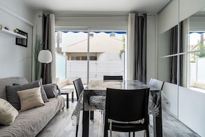 A modern living room with a sofa, a glass dining table, and chairs. There is a large window with curtains, and a view of a building outside. - Le Royal Studio adorable avec terrasse (Cannes)