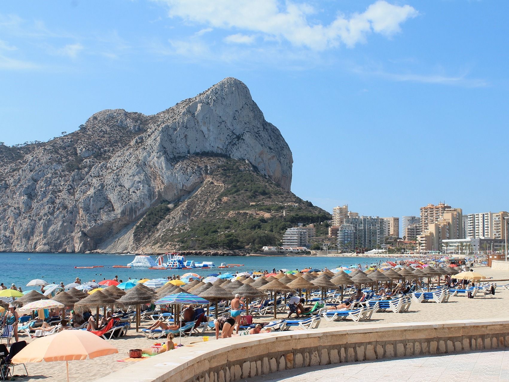 A beach with sun loungers and umbrellas, a rocky mountain in the background, and a cityscape with buildings.