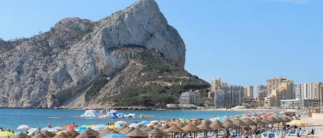 A beach with sun loungers and umbrellas, a rocky mountain in the background, and a cityscape with buildings.