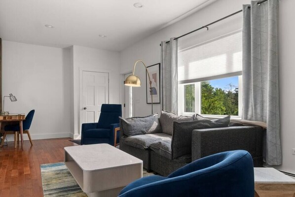 A modern living room with a sofa, a coffee table, and a blue chair. There is a dining area with a table and chairs. A large window with curtains is visible. - Hilltop 5-BR Carriage House 4-Min to Okemo (Ludlow)