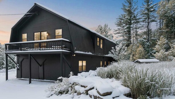 Una casa de dos pisos de color oscuro con un balcón, rodeada de árboles nevados y un paisaje cubierto de nieve.