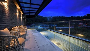 Un espace piscine aménagé sur le toit, avec des fauteuils en osier et une balustrade en verre donnant sur une vue panoramique de la ville au crépuscule.
