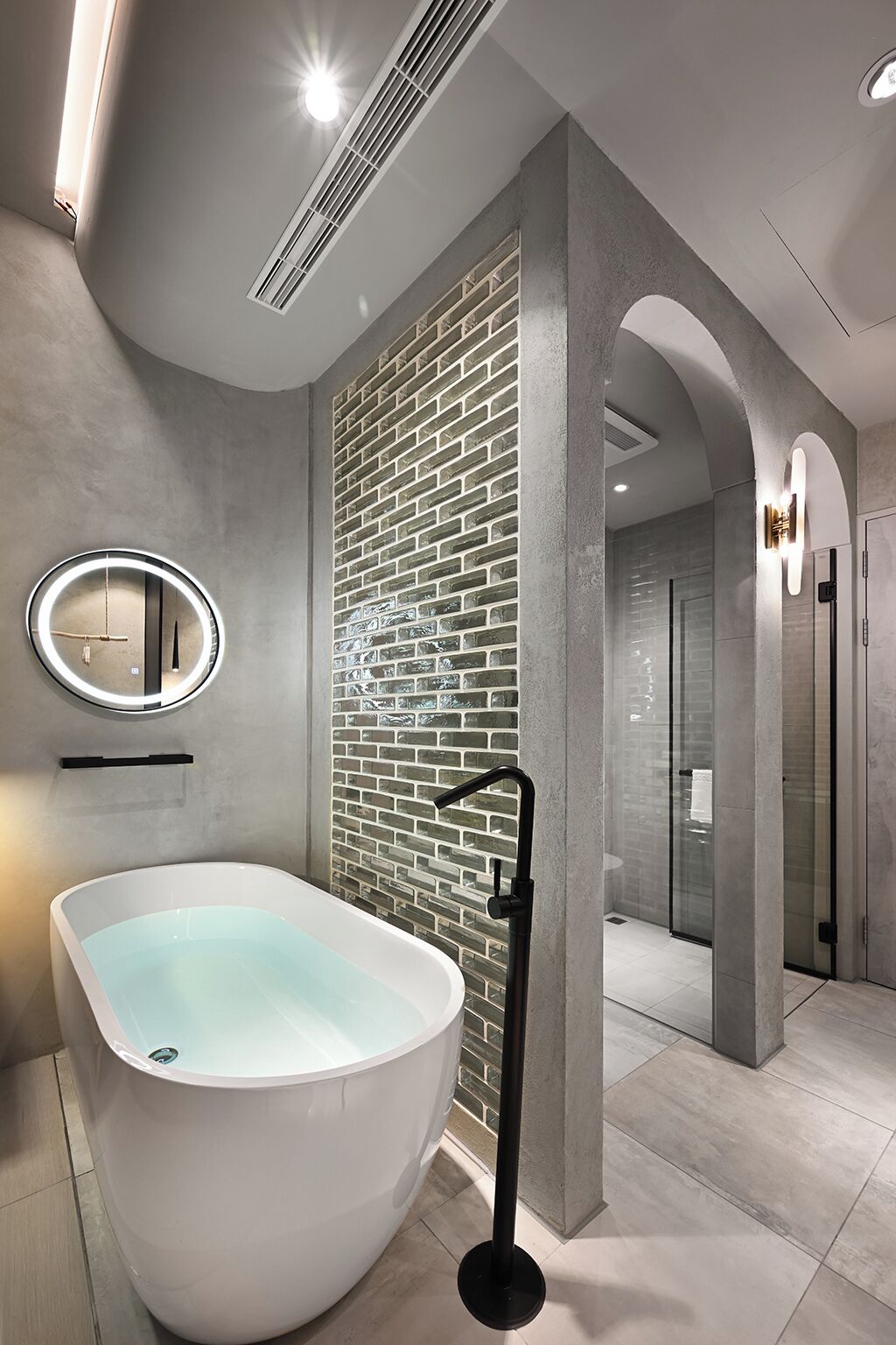 A modern bathroom with a freestanding bathtub, a round mirror with integrated lighting, and a brick-patterned accent wall.