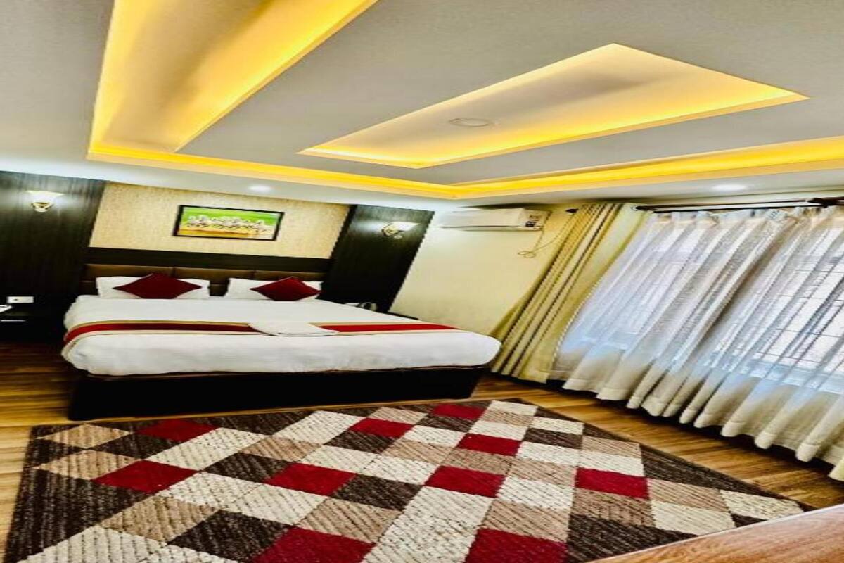 A hotel room with a bed, a patterned rug, a ceiling with recessed lighting, and a window with curtains.