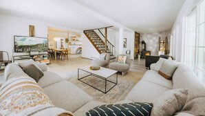 A spacious living room with a large sofa, a coffee table, and a TV. There is a staircase leading to an upper level.