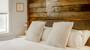 A bed with white bedding and decorative pillows, a wooden headboard, a bedside table with a lamp, and a framed picture on the wall.