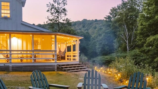 A cozy outdoor seating area with Adirondack chairs and a fire pit, surrounded by a house with large windows and a porch.