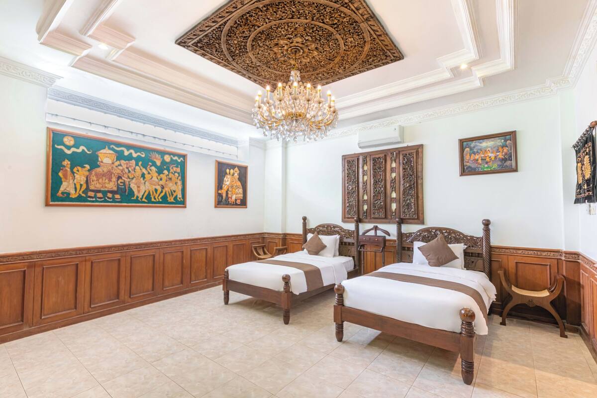 Habitación de hotel con dos camas de madera, una gran araña decorativa y arte mural tradicional.