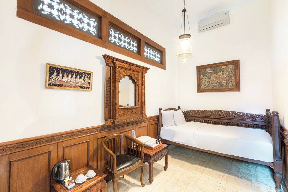 Una habitación con una cama de madera, una silla de madera, una mesita con una tetera y dos marcos de cuadros en la pared.