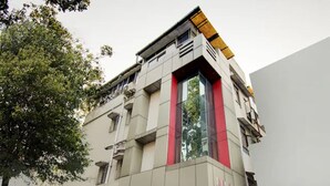 Un immeuble à plusieurs étages doté d’une architecture moderne, caractérisé par une palette de couleurs dominante en rouge et gris, de grandes fenêtres et un toit plat surmonté d’un auvent jaune.