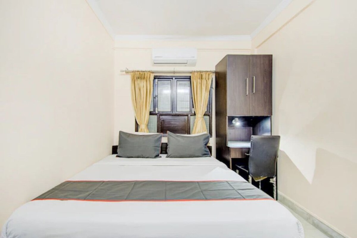 A bedroom with a bed, two pillows, a desk with a chair, a wardrobe, and a window with curtains.