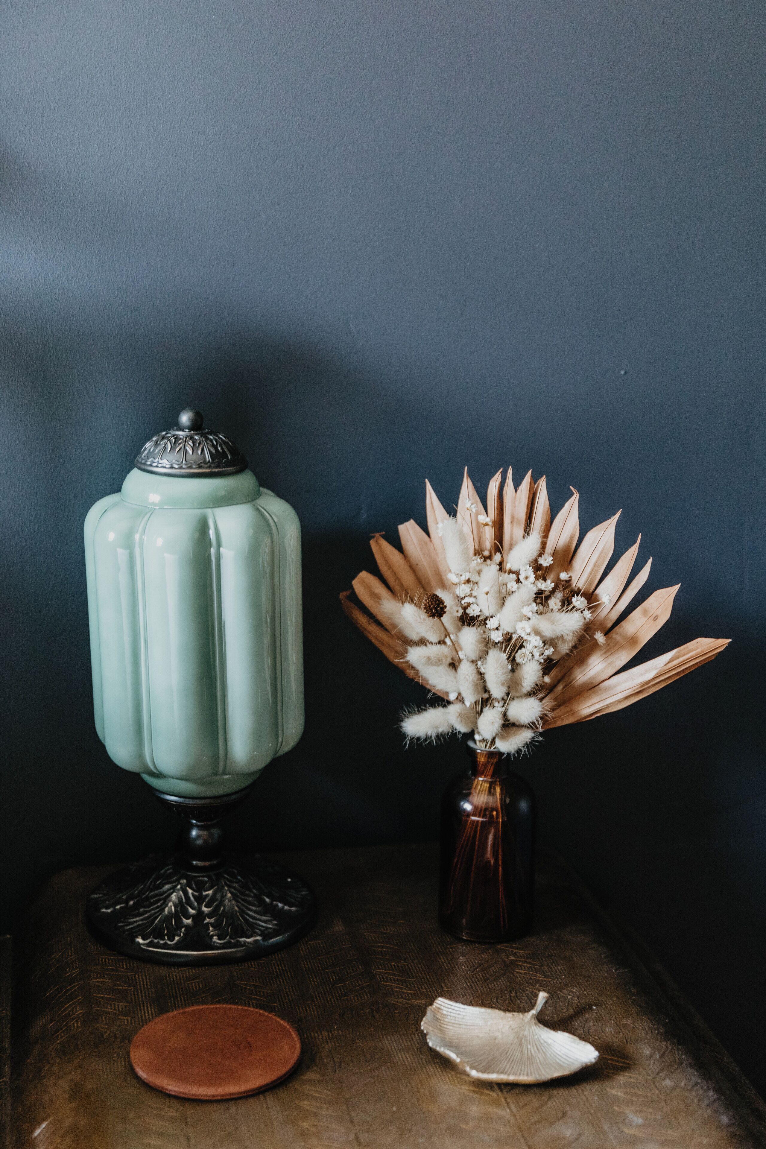 A vintage-style ceramic jar with a lid, a small vase with dried flowers, and a single seashell on a wooden surface against a dark wall.