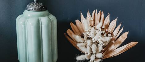 A vintage-style ceramic jar with a lid, a small vase with dried flowers, and a single seashell on a wooden surface against a dark wall.
