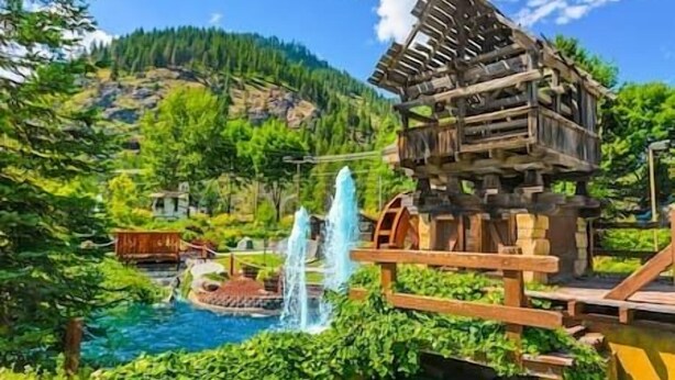 A rustic wooden cabin with a wooden bridge over a pool, surrounded by lush greenery and a mountainous backdrop. - Icicle Village 2-Bedroom w/ Pool (Leavenworth)