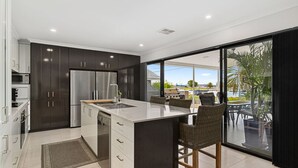 A modern kitchen with dark cabinetry, a stainless steel refrigerator, and a large island with bar stools. - Water Lovers Haven in Mandurah (Halls Head)