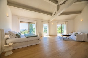 Un dormitorio amplio con una cama grande, una zona de estar con una mesa y un sofá. Amplias ventanas ofrecen luz natural y vistas al exterior.