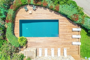 Vista aérea de una piscina rectangular con cubierta de madera, rodeada de exuberante vegetación y un muro de piedra.