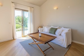 Una habitación con una mesa de centro de madera, un sofá blanco con cojines y vistas a través de un ventanal.