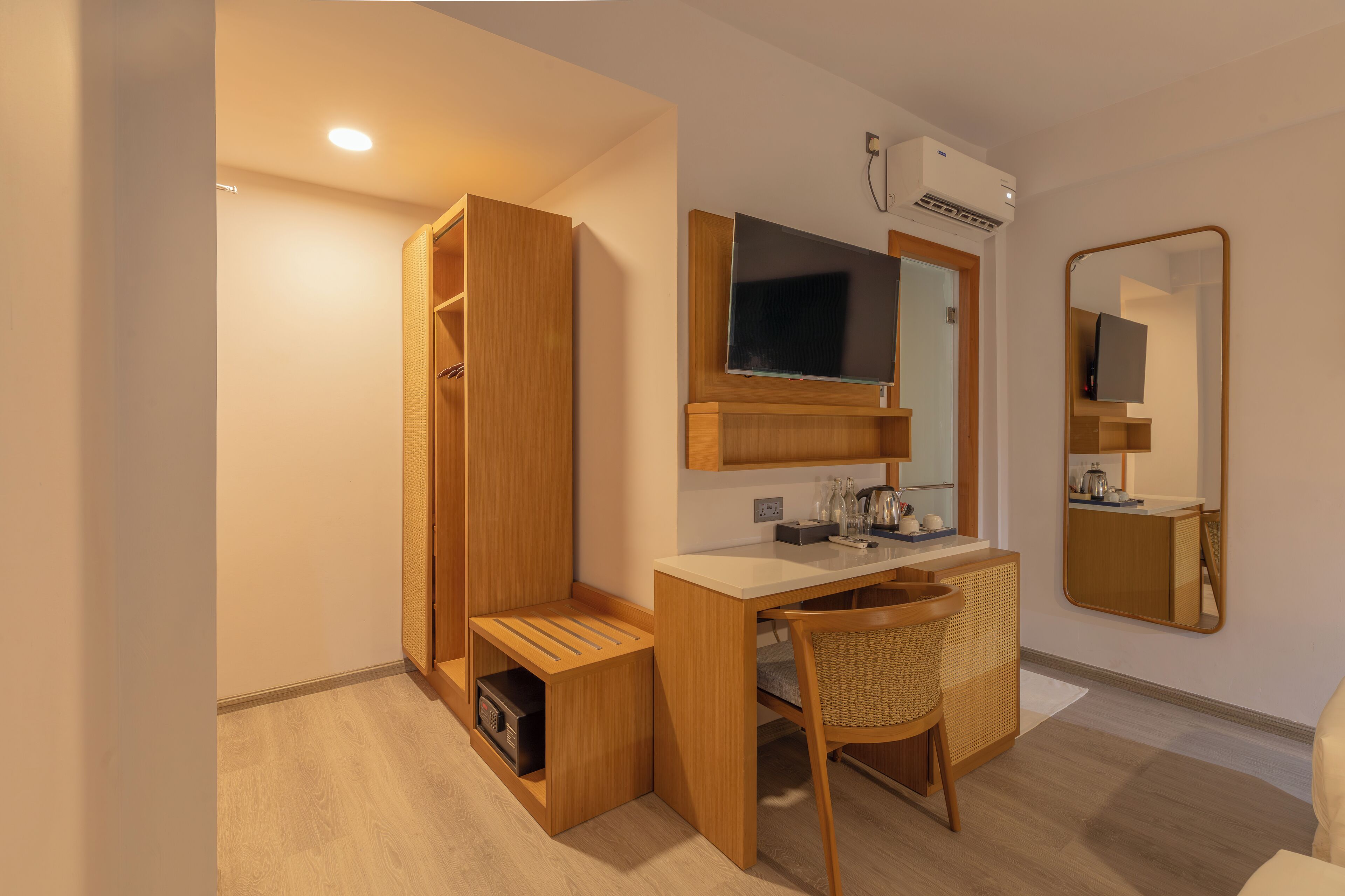 A compact room with a wooden desk, a chair, a wardrobe, a mirror, and a flat-screen TV.