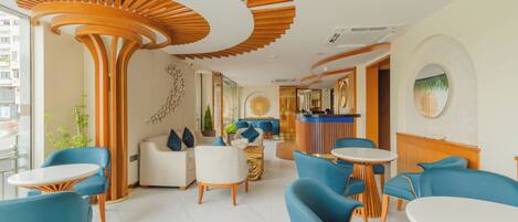 A modern hotel lobby with a central seating area, a reception desk, and a large mirror on the wall.