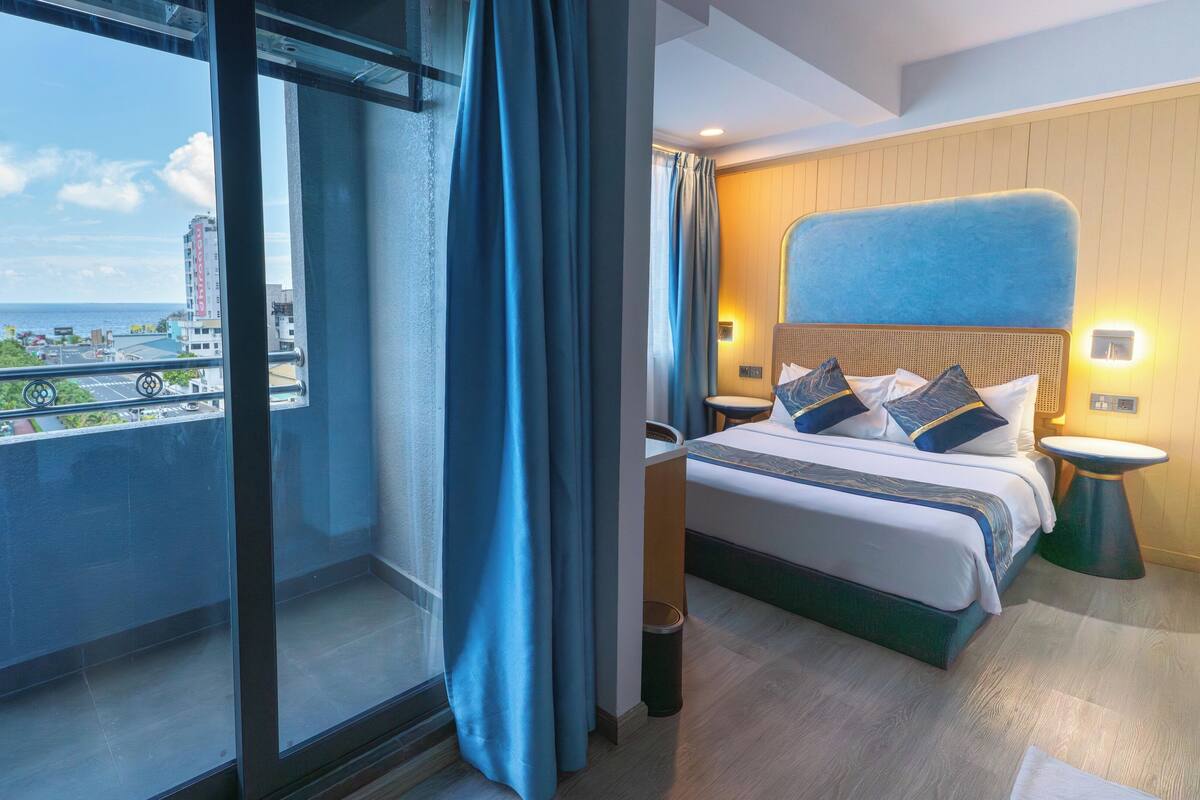 A hotel room with a large window, a bed with pillows, a bedside table, and a wall-mounted light.
