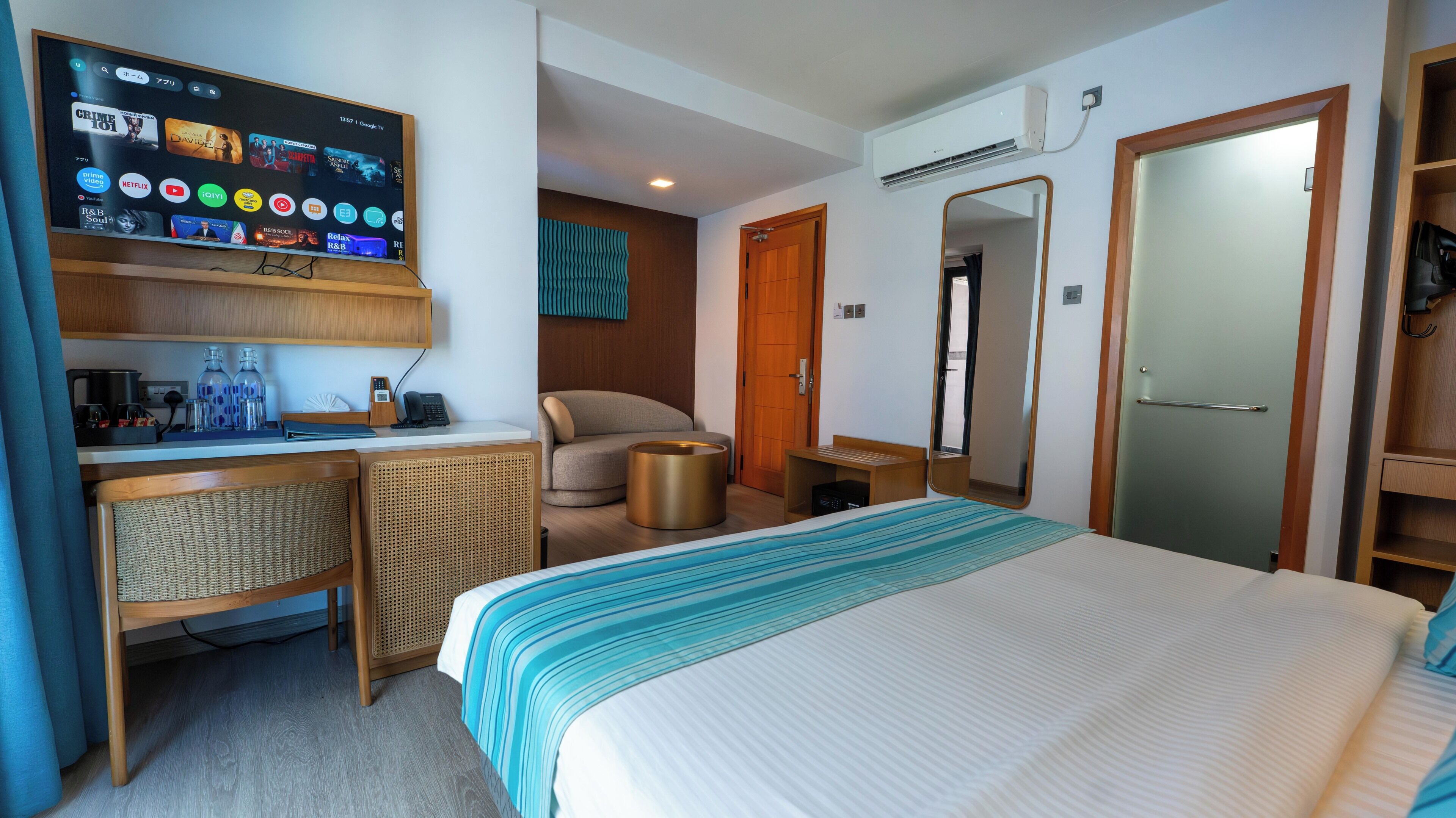 A hotel room with a bed, a desk with a computer, a chair, a small table, a mirror, and an air conditioning unit.