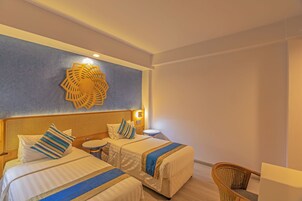 A hotel room with two beds, a round table, a wicker chair, and a decorative wall piece.