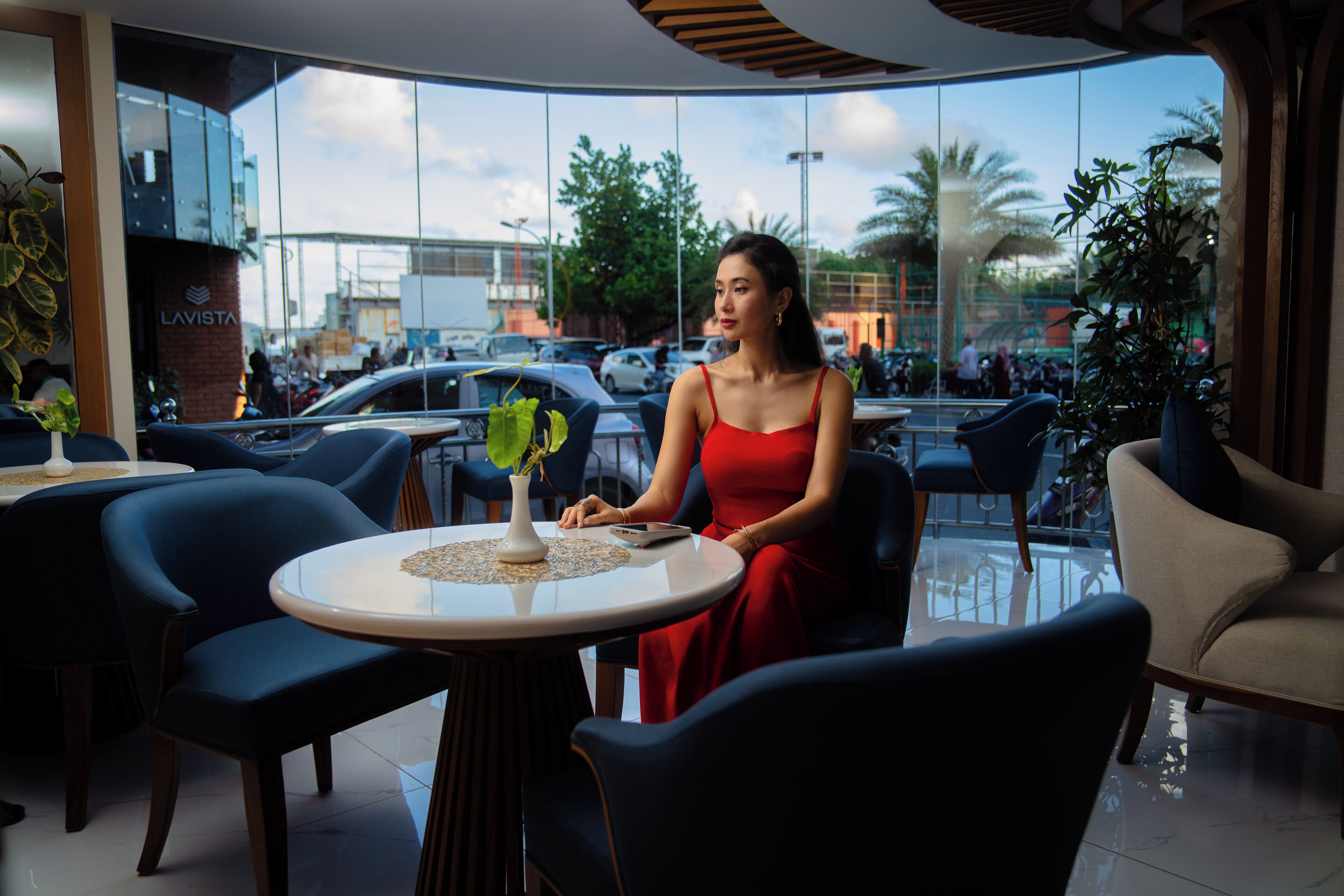 A woman in a red dress sitting at a round table with a vase of green plants, in a modern indoor space with large windows and blue chairs.
