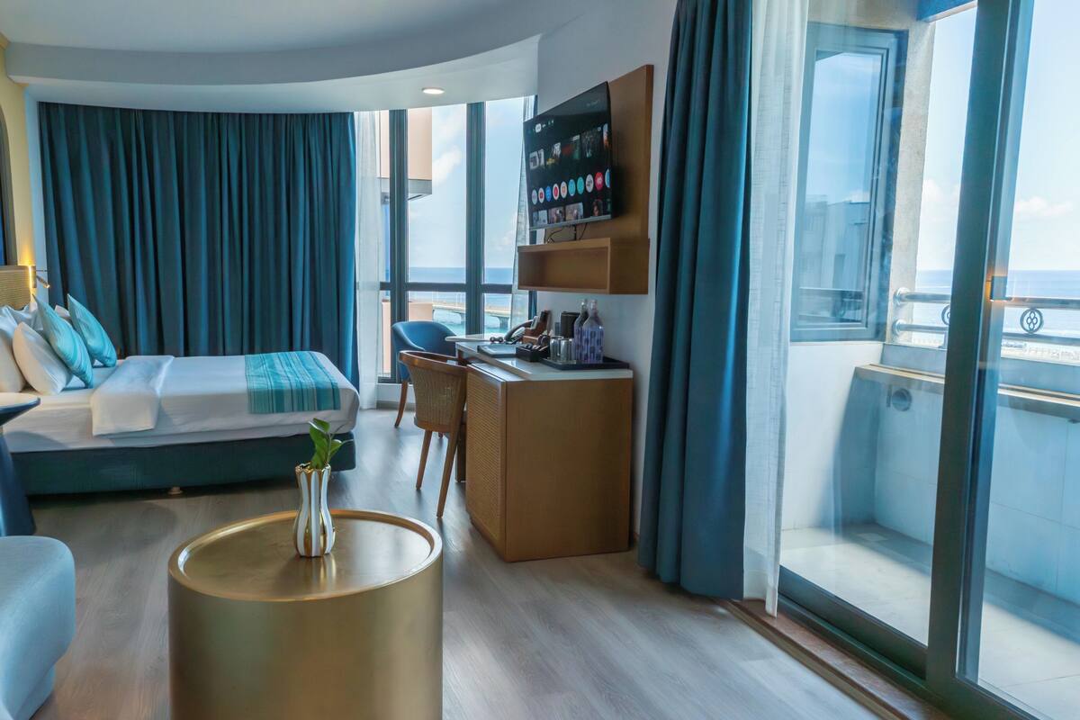 A modern hotel room with a bed, desk, chair, and a view of the sea.