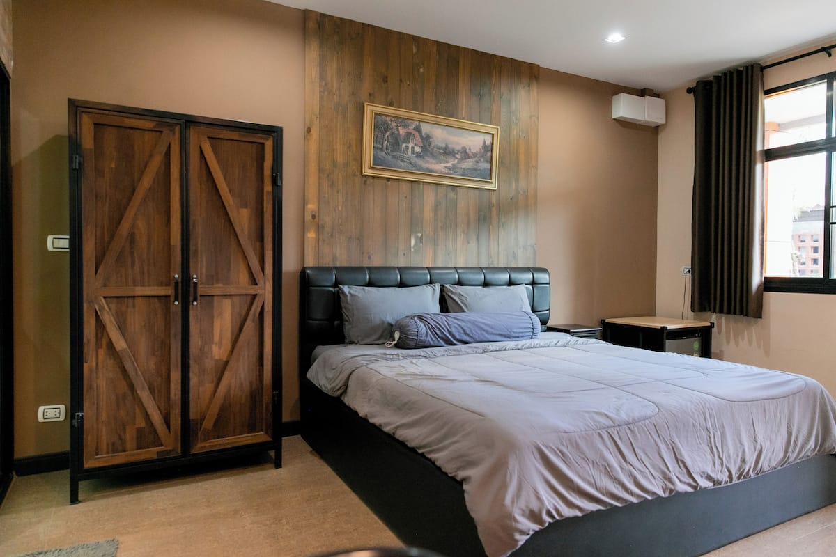 A bedroom with a large bed, a wooden wardrobe, a small bedside table, and a painting on the wall.