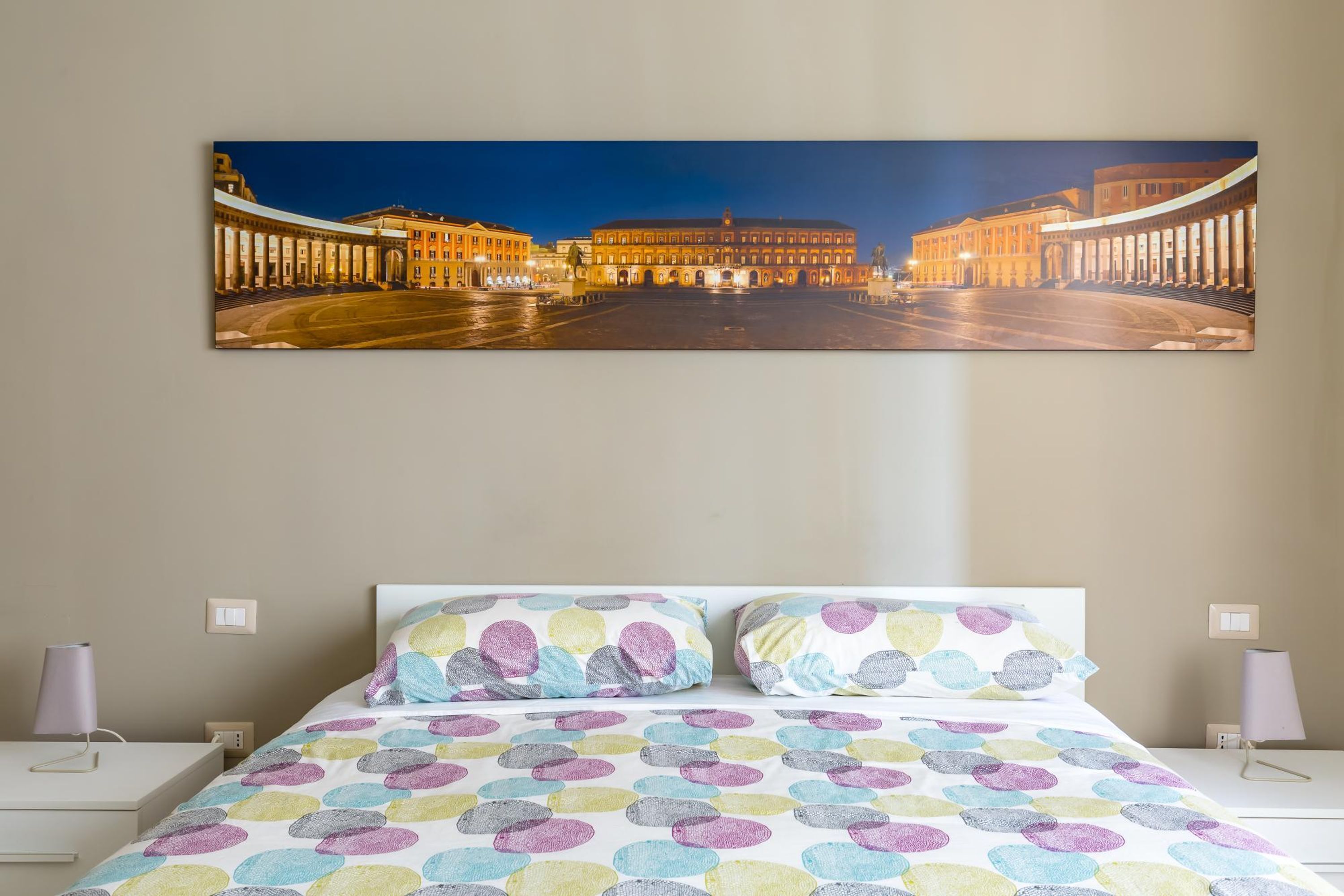 Ett hotellrum med en stor, inramad tavla som föreställer en stadsbild, en säng med färgglatt, cirkelformat sängklädnad och sänglampor.
