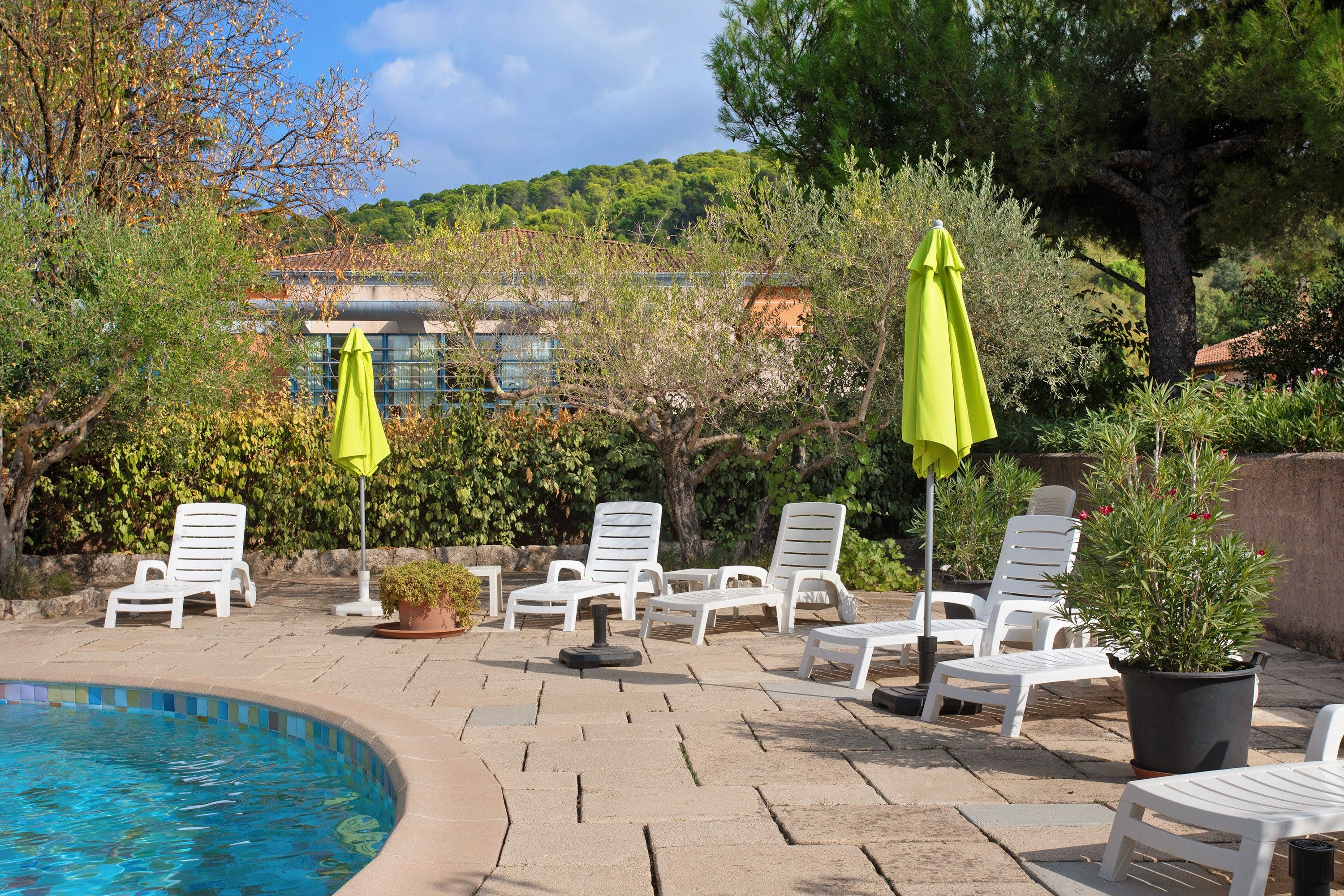 A poolside area with lounge chairs and umbrellas, surrounded by greenery and a building in the background.