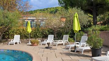 A poolside area with lounge chairs and umbrellas, surrounded by greenery and a building in the background.