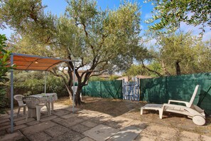 An outdoor patio with a table and chairs, a covered seating area, and a green fence. - Studio Apartment 'Meublé' with Shared Pool, Private Garden and Wi-Fi (Lorgues)