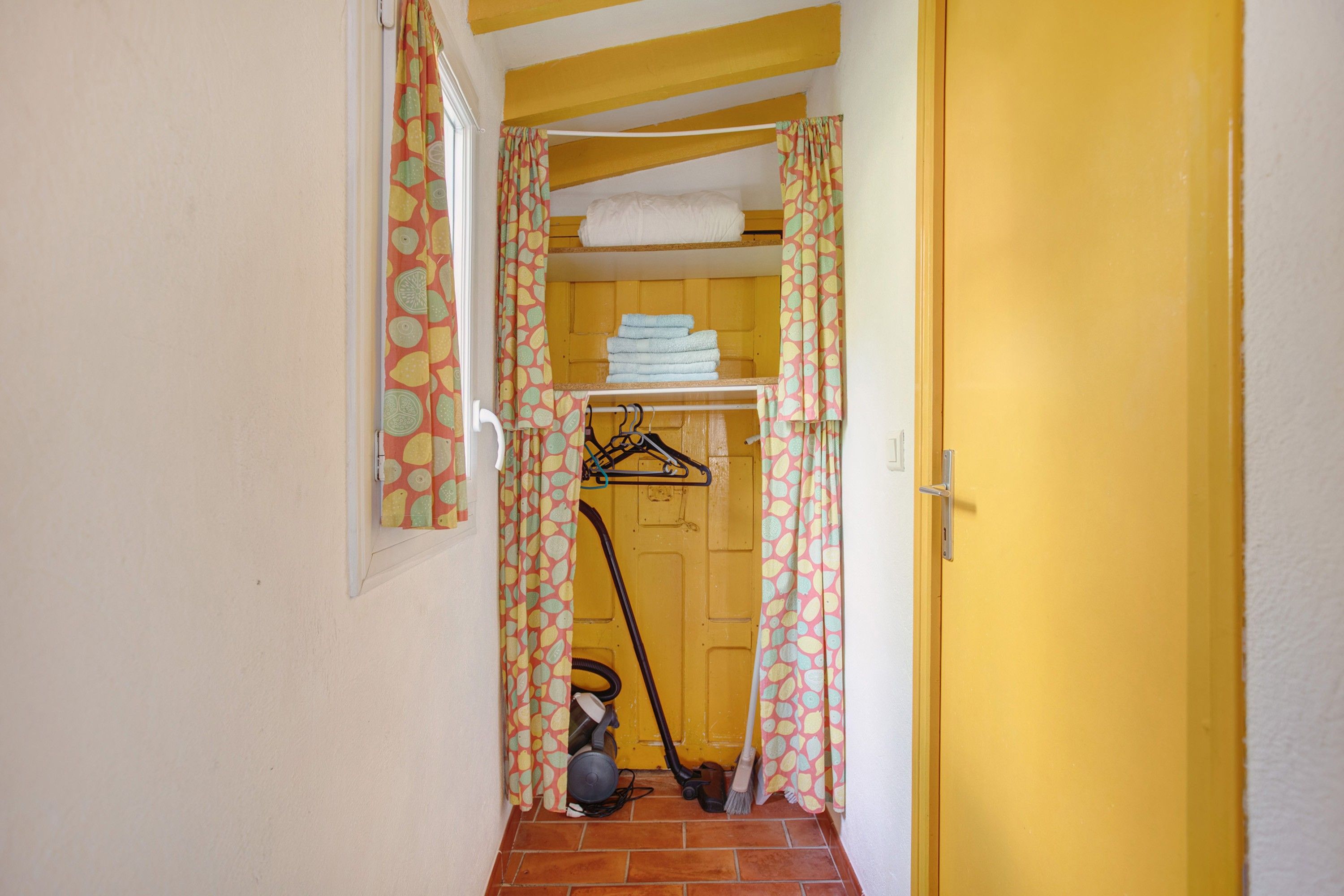 Un pasillo estrecho con una puerta amarilla, un área de lavandería con ropa colgada y una aspiradora.