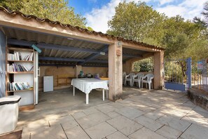 A covered outdoor area with a table and chairs, a bookshelf, and a refrigerator.