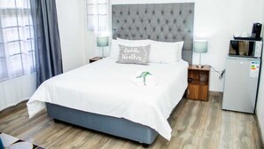 A neatly made bed with a grey headboard, two bedside tables with lamps, a small wooden nightstand, and a refrigerator. - Erima lodge (Johannesburg)