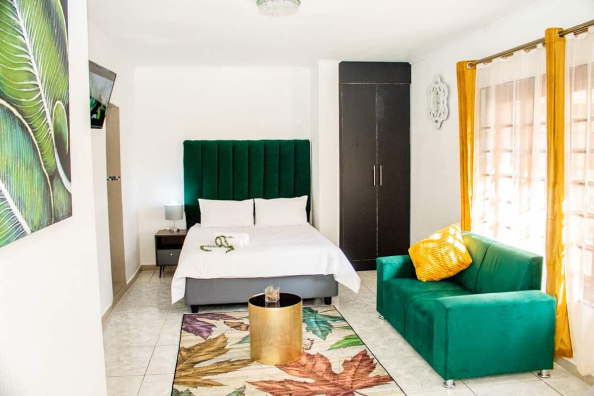 Une chambre d’hôtel avec un lit, un canapé vert, une armoire et une grande fenêtre avec des rideaux jaunes.