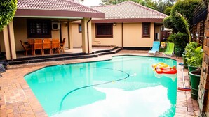 A swimming pool with a tiled deck, surrounded by a house with a tiled roof and a covered patio area with wooden furniture. - Erima lodge (Johannesburg)