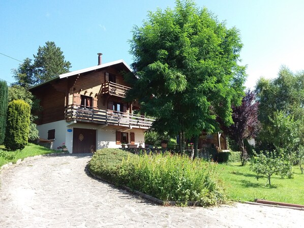 A two-story wooden house with a balcony, surrounded by greenery and a cobblestone path.