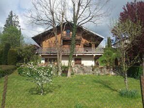 A wooden chalet with a balcony, surrounded by greenery and a stone wall.