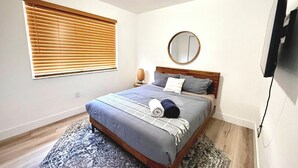 Et soveværelse med en seng, et rundt spejl, et sengebord med en lampe, et vindue med persienner og et fjernsyn monteret på væggen.