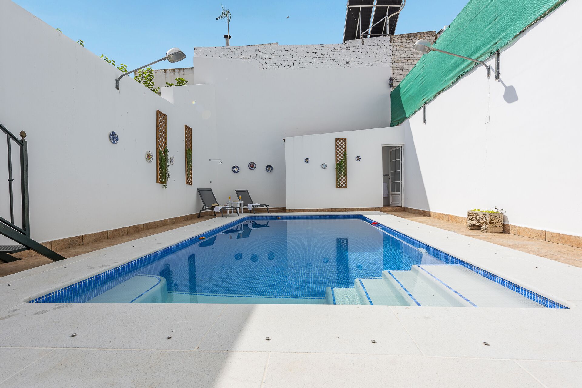 A swimming pool with a tiled deck, a covered area with a green tarp, and a door leading to a building.