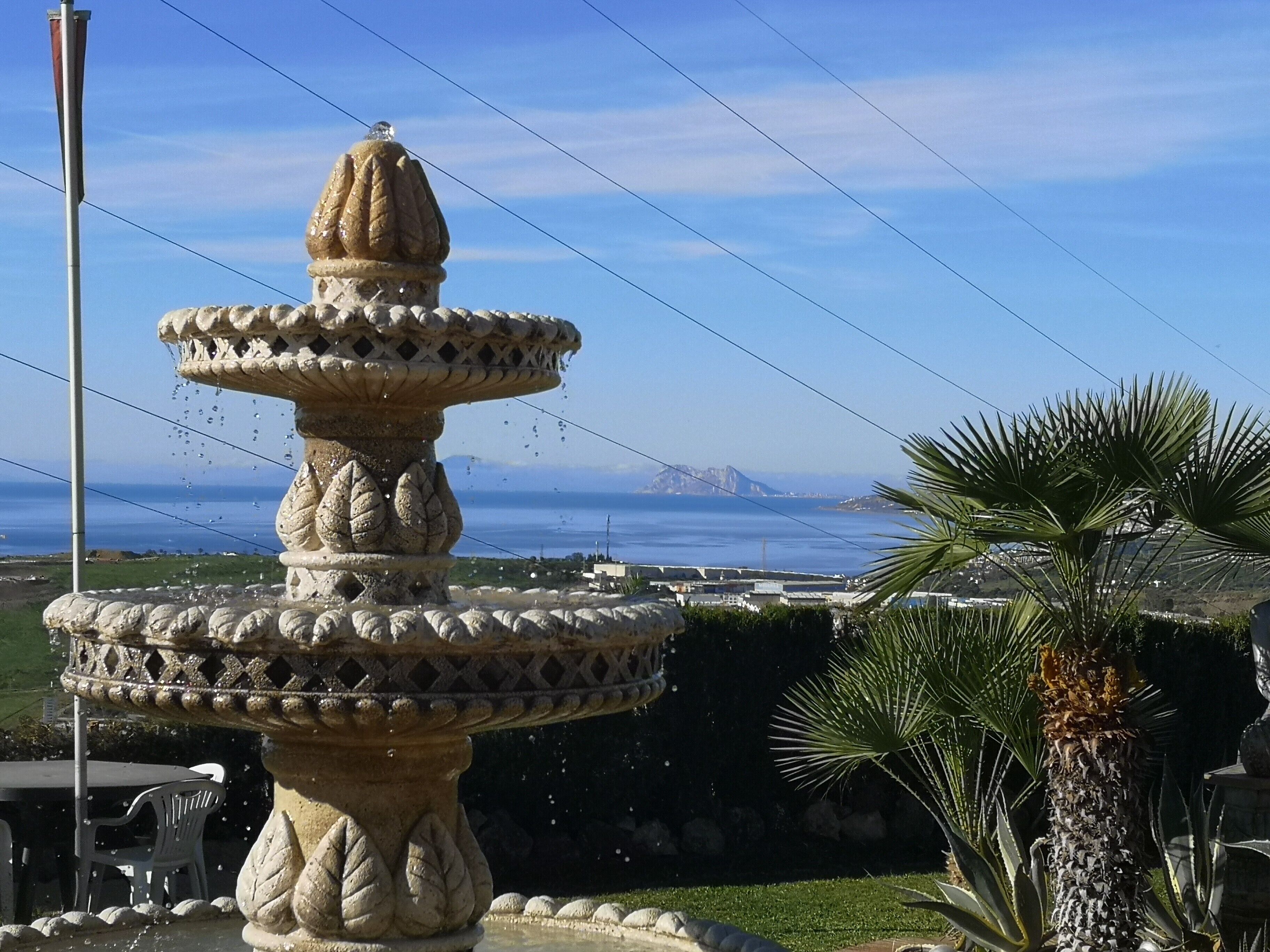 A stone fountain with a tiered design, surrounded by greenery and overlooking a coastal view.