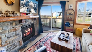 Una sala de estar acogedora con chimenea de piedra, un televisor plano grande y vistas a una montaña nevada a través de un ventanal corredera.