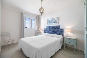 Un dormitorio con cama, una silla, una mesita de noche, una lámpara, una ventana y una obra de arte colgada en la pared.