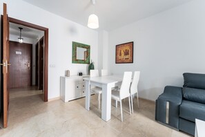 Ein modernes Wohnzimmer mit einem weißen Esstisch, weißen Stühlen, einem grauen Sofa, einer Holztür und einem gerahmten Kunstwerk an der Wand.