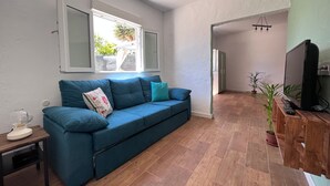 Ein Zimmer mit einem blauen Sofa, einem Fernseher auf einem Holztisch, einem Fenster mit Vorhängen und einer Tür zu einem weiteren Raum.